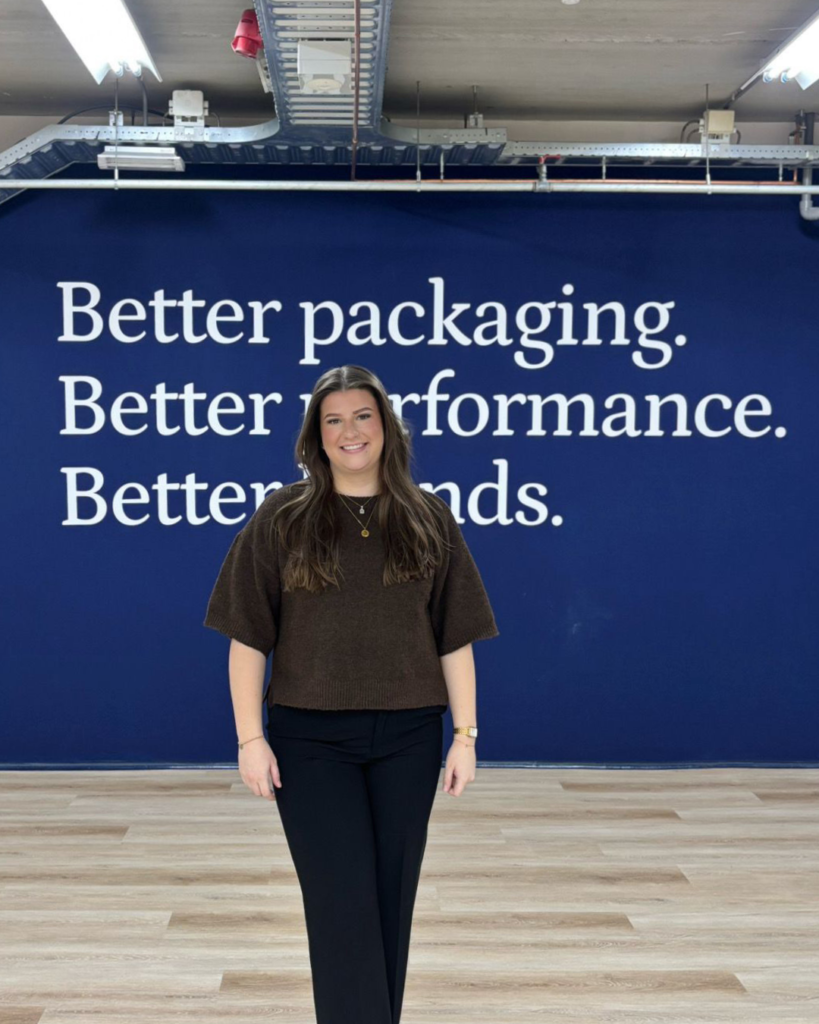 De baan van Lotte - Eshuis - BIND Vrouw staat glimlachend rechtop in een ruimte met een houten vloer, voor een blauwe muur met de witte tekst “Better packaging. Better performance. Better brands.” Ze draagt een donkerbruine trui, zwarte broek en sneakers.